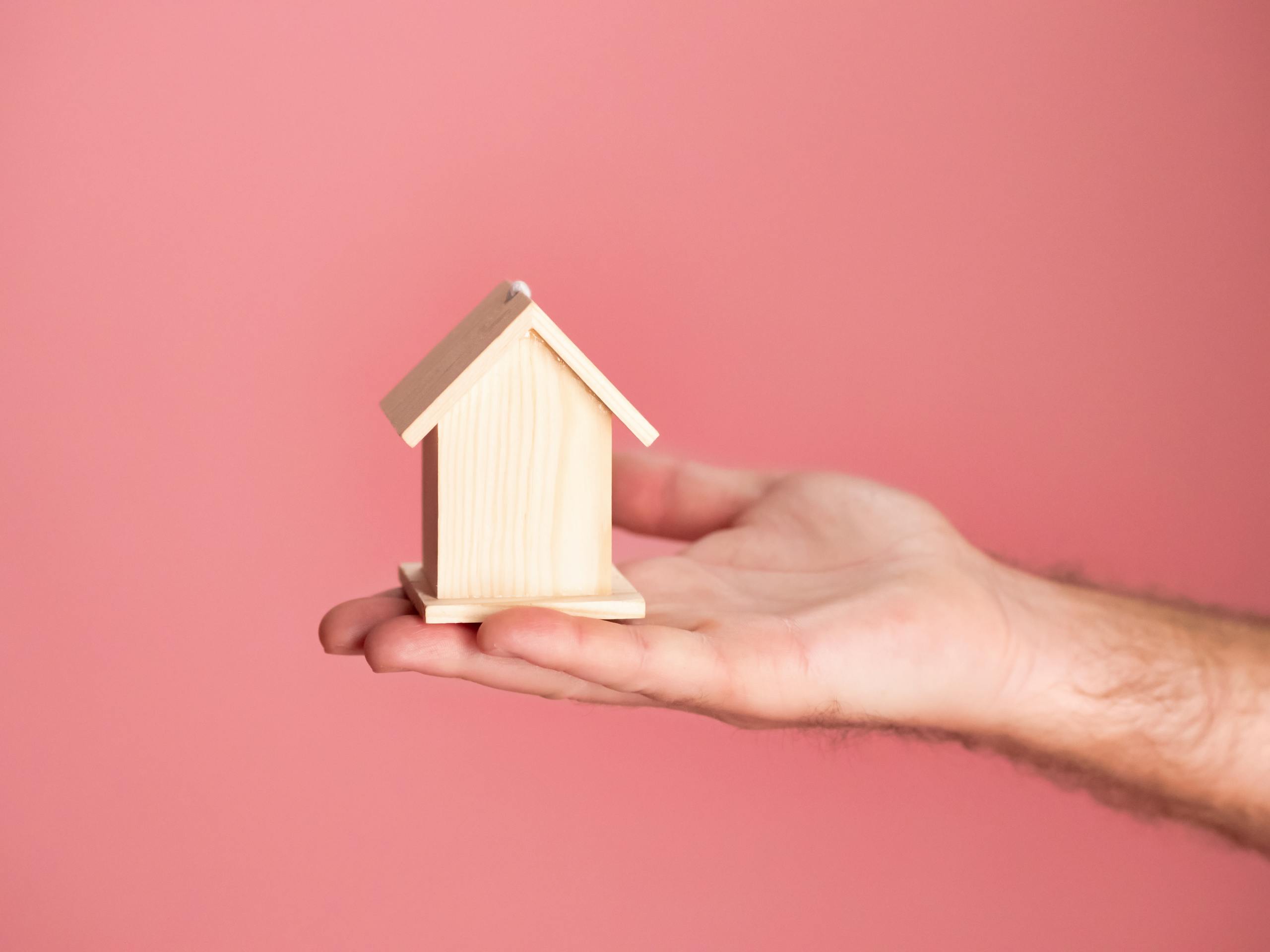 Hand holding a small wooden house model against a soft pink background, symbolizing real estate and property investment.